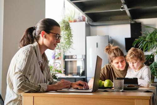 A light-skinned woman sits working a laptop with her partner and child sitting in the background.