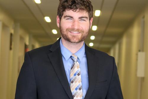 A white, masculine-presenting individual with a brown beard that covers the majority of their face is dressed in a suit and tie and smiles at the camera. They are located in a hallway within the College of Education at the University of Massachusetts Amherst. 