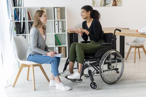 A medium-dark skinned woman sitting in a wheelchair chats with a light-skinned woman sitting in a chair.