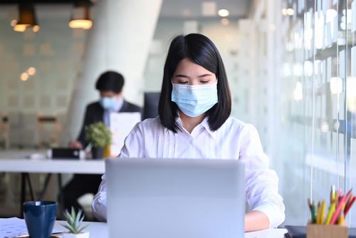A medium skinned woman with a mask on types on a laptop.