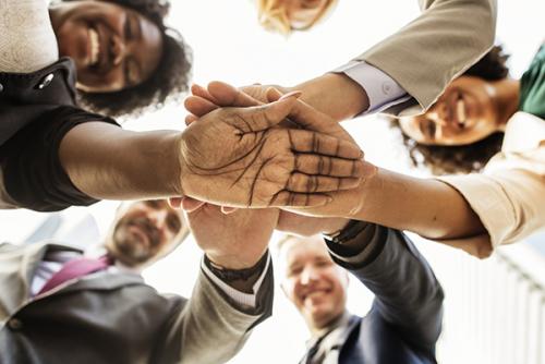 People with diverse skin tones look down at a camera with their hands stacked over each others' in a pact.