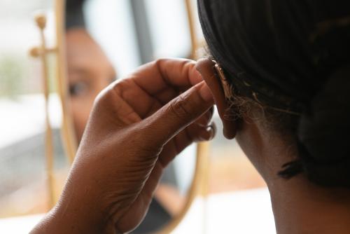 A dark-skinned woman fixes her hearing aid in a mirror.