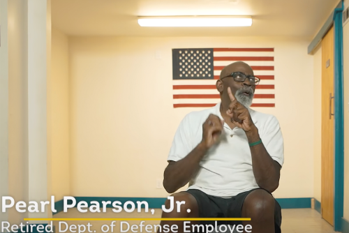 Pearl Pearson, Jr., a dark-skinned man sits in a chair signing with the American flag on a wall behind him.