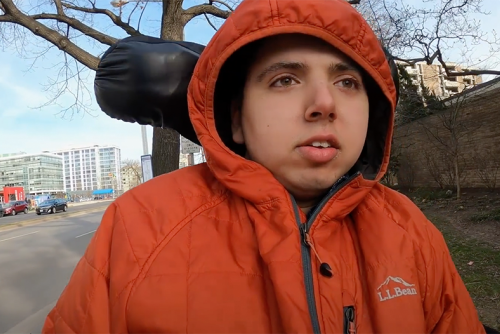 Samuel Habib, an olive skinned young man wears a winter jacket with the hood over his head as he rides down a sidewalk in his motorized wheelchair.