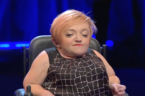 Stella Young, a light-skinned woman sits in a motorized wheelchair presenting on a stage.