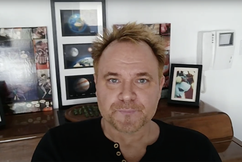 Mik Scarlet, a light-skinned man, sits with a table behind him displaying photos of planets and a woman.