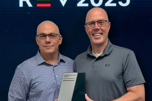 Two UMass employees standing side by side holding an award in front of a logo for company Jaggaer and a graphic logo for the REV 2025 conference