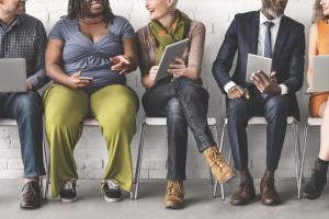 a diverse group of individuals in business attire sit in a row of chairs looking at their laptops and tablets