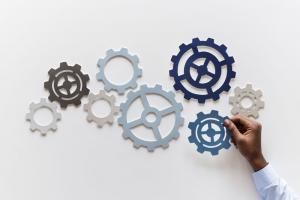 a dark skinned male hand in a blue shirt adds a gear to a collection of interconnected gears laid out on a white table 