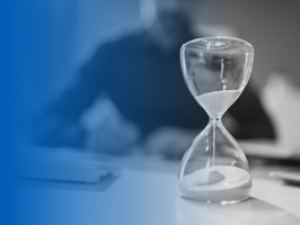 Glass hourglass with sand falling from top to bottom on top of desk with a person sitting in the background looking at papers.