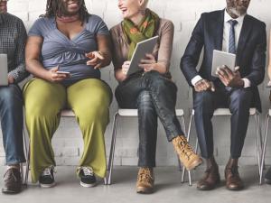 a diverse group of individuals in business attire sit in a row of chairs looking at their laptops and tablets