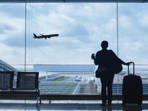  Person stands in front of a large window with a suitcase looking out at an airplane in the sky taking off at an airport