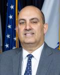 Stephen Zrike is smiling in front of an American flag and the Massachusetts state flag in the background wearing a light grey suit, white dress shirt and navy blue tie with designs