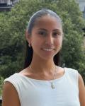 Student Trustee Ashley Chamu Charco is smiling, wearing a white short-sleeve shirt and a necklace, standing in front of a tree in the background