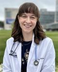 Student Trustee Jayne Vogelzang is smiling, wearing a white medical jacket with a black shirt underneath with flowers and a stethoscope around her neck, sitting near a grassy field in front of a campus background
