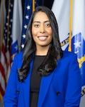 Juana Matias in a royal blue blazer and black shirt with American flag and Massachusetts state flagin background