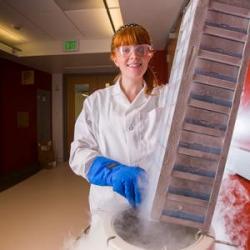 A light-skinned woman with red hair is dressed in lab attire with latex gloves. She holds a container of specimens. 