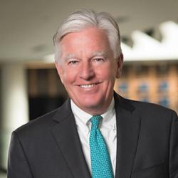 President Meehan, a man with medium-light skin, white hair, and a cleanly shaven face, smiles at the camera. He wears a dark gray suit coat, an aqua tie, and a white shirt.