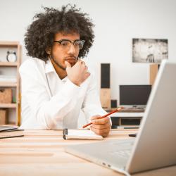 A dark-skinned man wearing glasses sits at a desk taking a note while looking at information on his computer screen.