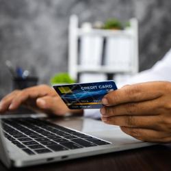 Medium skin toned hands work on a laptop, the left hand holding a credit card.