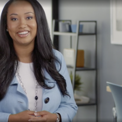 A woman smiles with her hands clasped. She has office shelving and pictures behind her.