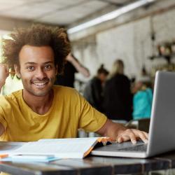 A dark-skinned young man wearing a long yellow sweatshirt sits at a coffee table with his laptop open and books on the table.