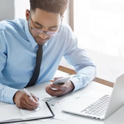 A dark-skinned man writes in a notebook with a computer to the side.