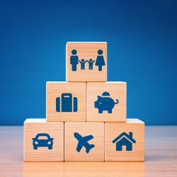Five wooden blocks are stacked. The bottom row has a car, plane, and house. The second row has a briefcase and piggy bank. The top row has a family.