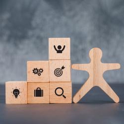 Three rows of wooden blocks. The bottom row has a light bulb, briefcase, and search icon. The second row has gears and a target. The top row is a person cheering.