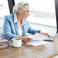 An light-skinned woman with white-gray hair sits at a desk working on a computer.