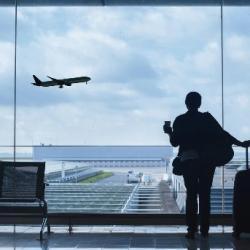 A person stands in an airport looking out the window at a plane taking off.