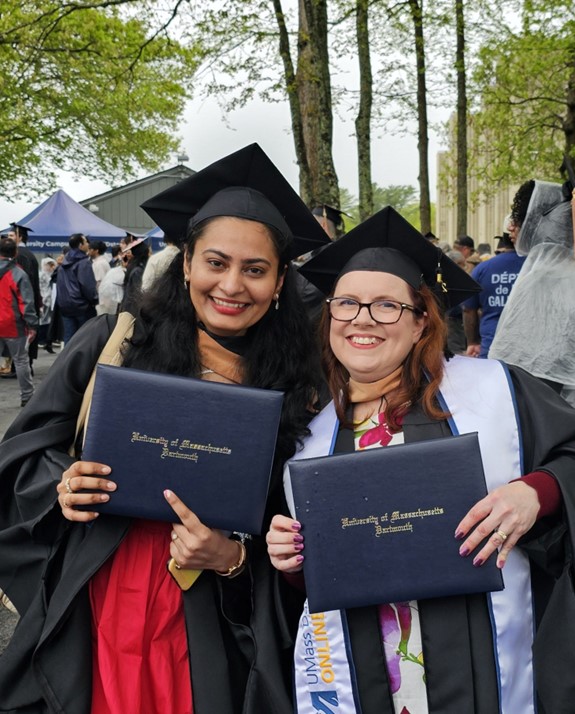 Two women stand together under trees amongst a crowd of graduate students and their families. They wear black graduation caps and gowns over brightly colored dresses and smile widely while displaying navy blue diploma jackets with embossed gold lettering reading "University of Massachusetts Dartmouth". The woman on the right is also wearing a UMass Dartmouth Online and Continuing Education graduation stole. The weather is grey and several people in the crowd are wearing rain ponchos. 